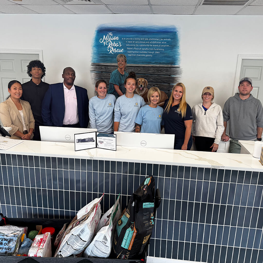 A group photo of the Reba's team celebrating Meagan Goins' Hometown Hero's Award. The background includes the mural, an award on the counter and bags of donated food.