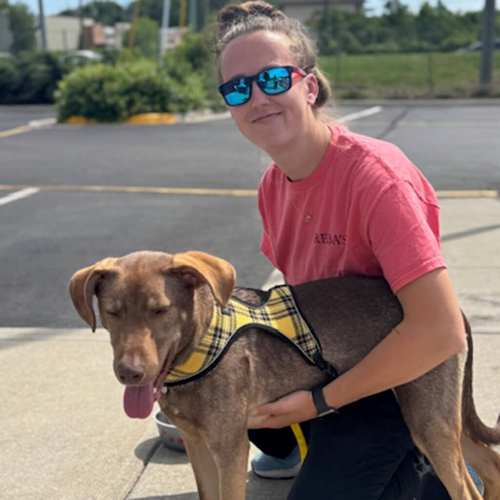Team member Hannah in red t-shirt has her arm around a brown dog. Dog has a yellow and black plaid harness on and is panting.