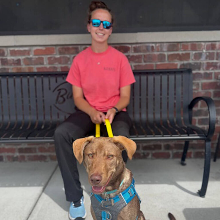 Team member Hannah is sitting on a bench outside Bubbas 33. A brown dog is sitting in front of her with a yellow leash.
