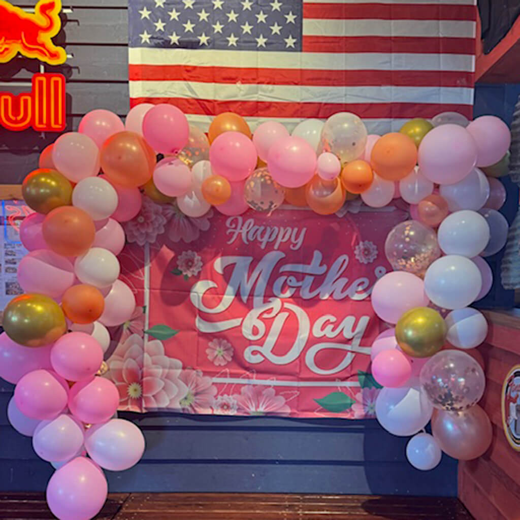 Pink, orange, gold, and white balloons surround a pink Happy Mother's Day sign. The American flag is above the balloons on a blue wall.