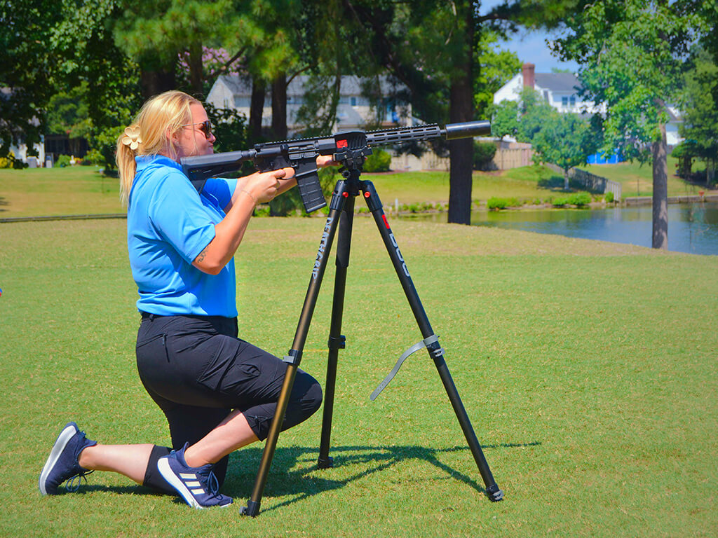 Meagan Goins is on one knee shooting the AR15 golf ball launcher. the background is the golf greens, water, trees and houses.