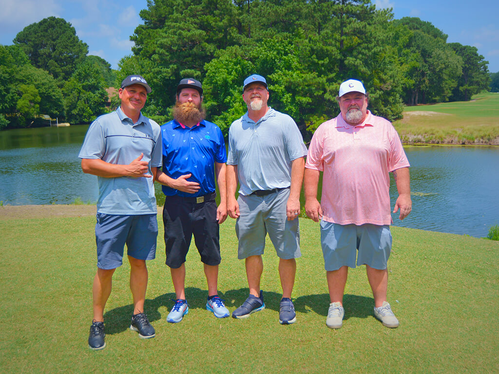 This was team 20. A group of four men standing on the green by the water with trees behind them.