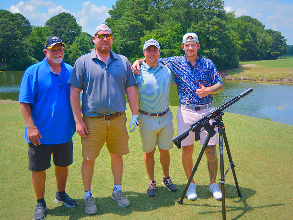 This was team 30. A group of four men standing on the green by the water with trees behind them. One man has his arm around the other guy giving a thumbs up and the launcher is in front of them.