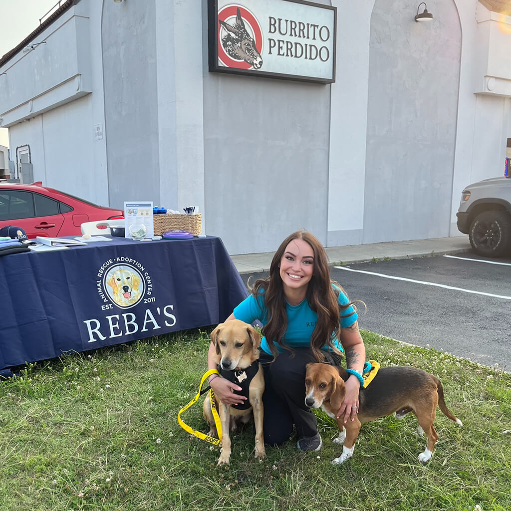 Lady in blue shirt kneeling down with two dogs in front of Burrito Perdido building.