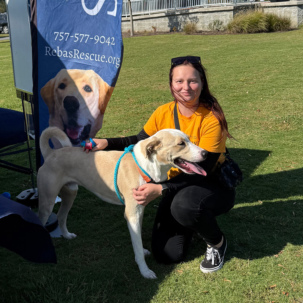 A volunteer in an orange t-shirt poses with a tan dog that is up for adoption.