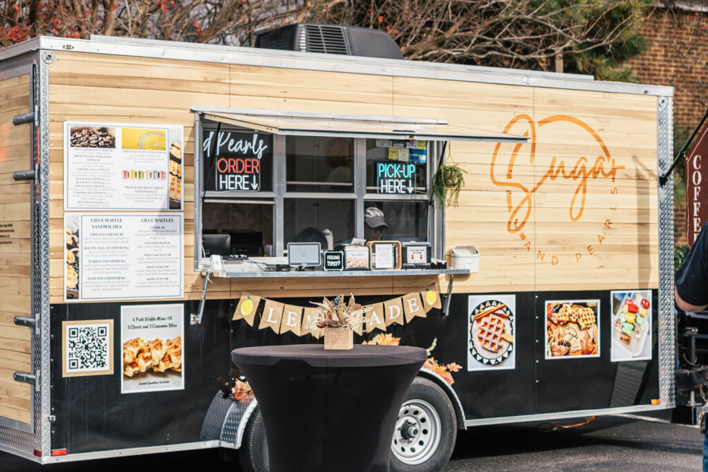Sugar and Pearls VB Food Truck. The outside of the truck is light colored wood and a black bottom with neon signs in the window.